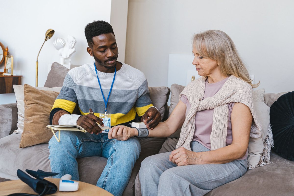 Care visitor in a lanyard recording a blood pressure reading for an older woman on a sofa