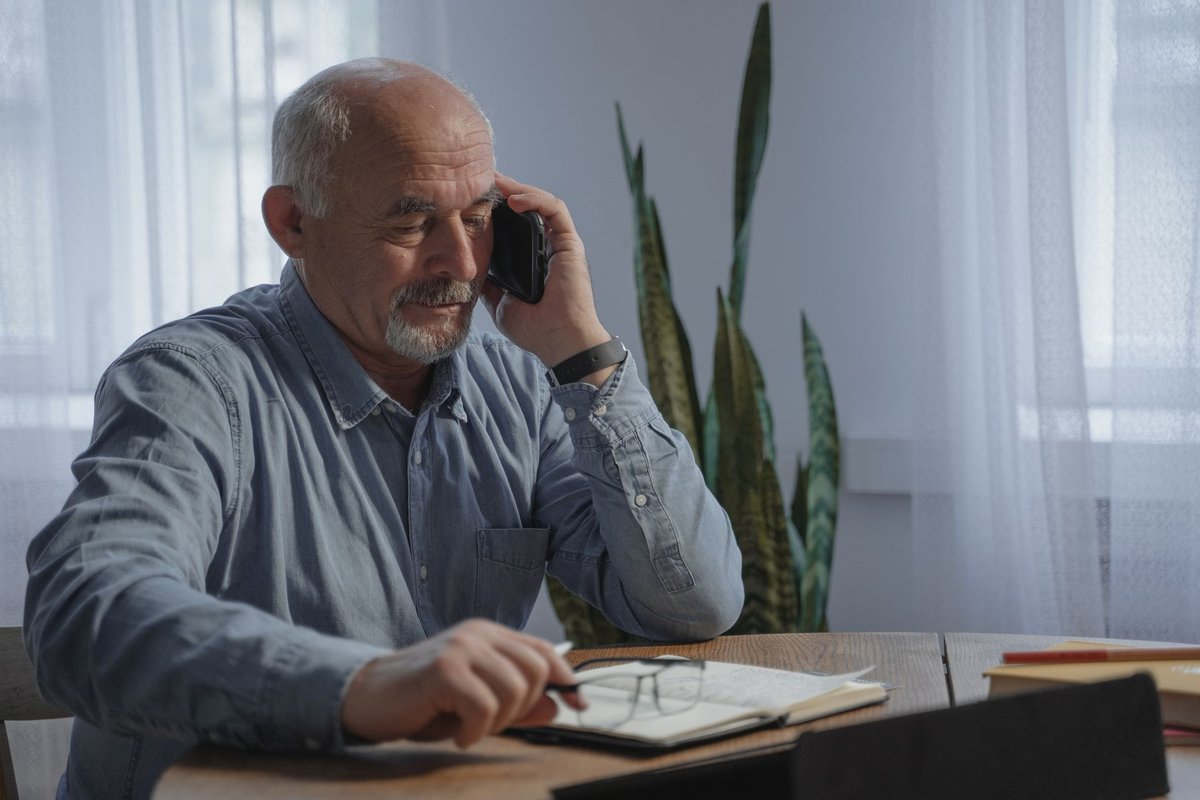 Older man on a phone call taking notes in a notebook at a wooden table