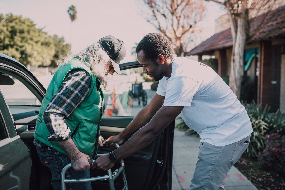 Helper steadying an older man with a walker as he steps out of a car