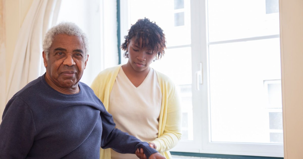 Caregiver supporting an older man's arm as he stands beside a bright window