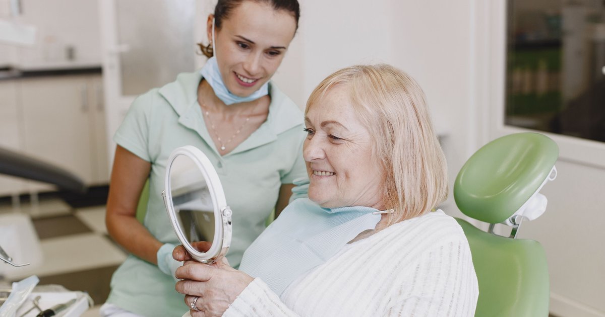 Dental hygienist smiling beside an older woman who holds a hand mirror in the dental chair