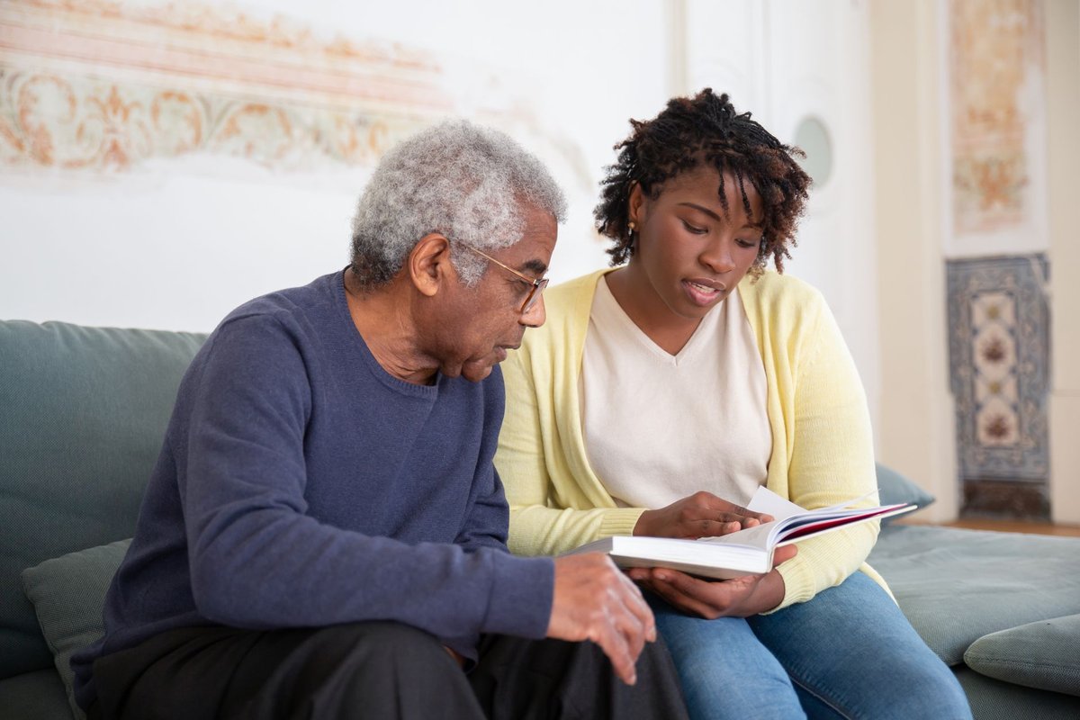 Older man and a caregiver reading a book together on a sofa at home