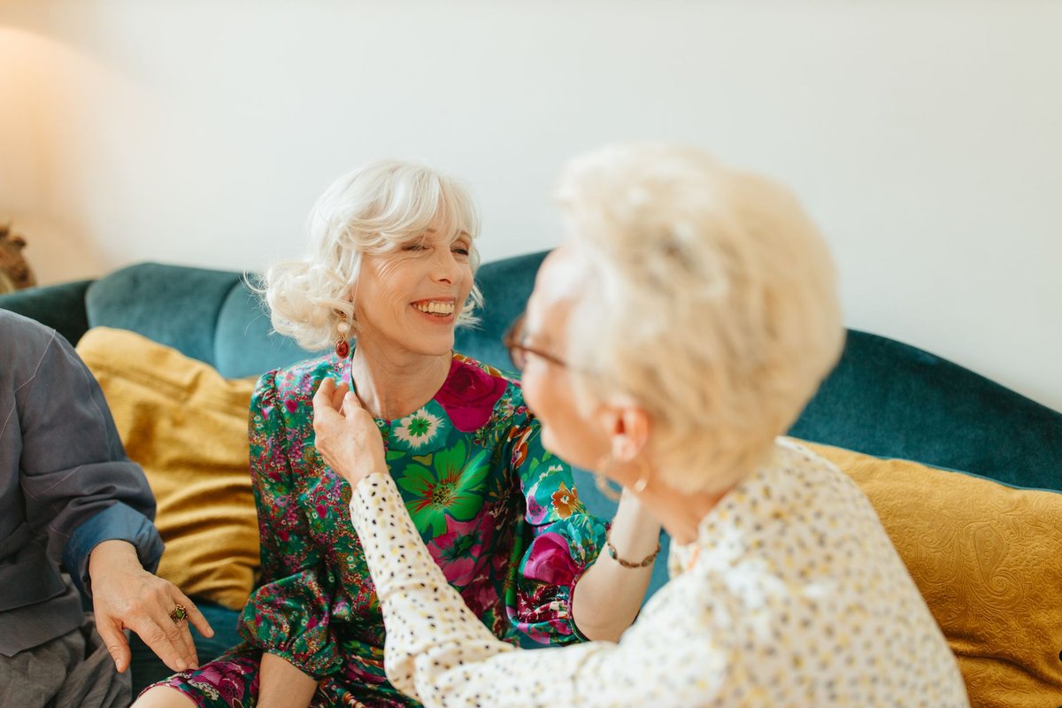 Two older women laughing together on a sofa as one admires the other's earring