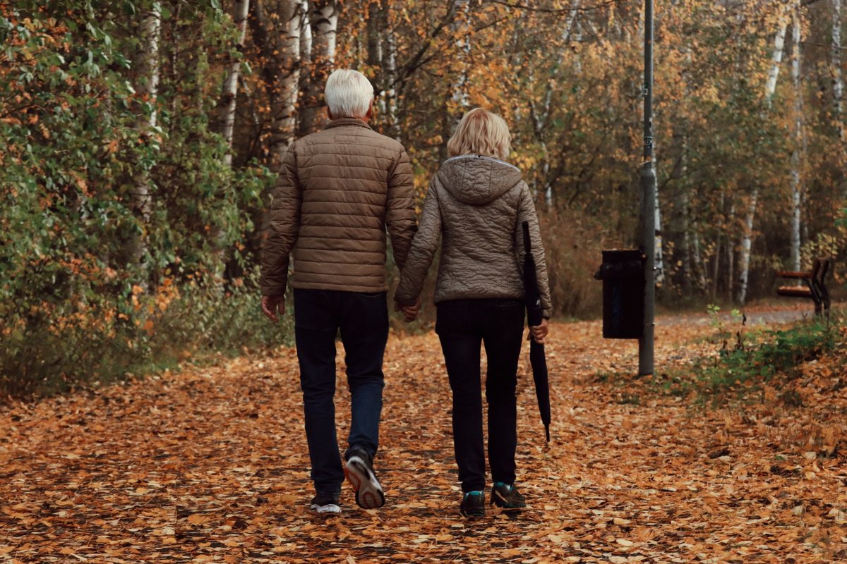 Older couple walking hand in hand along a leaf-covered park path in autumn