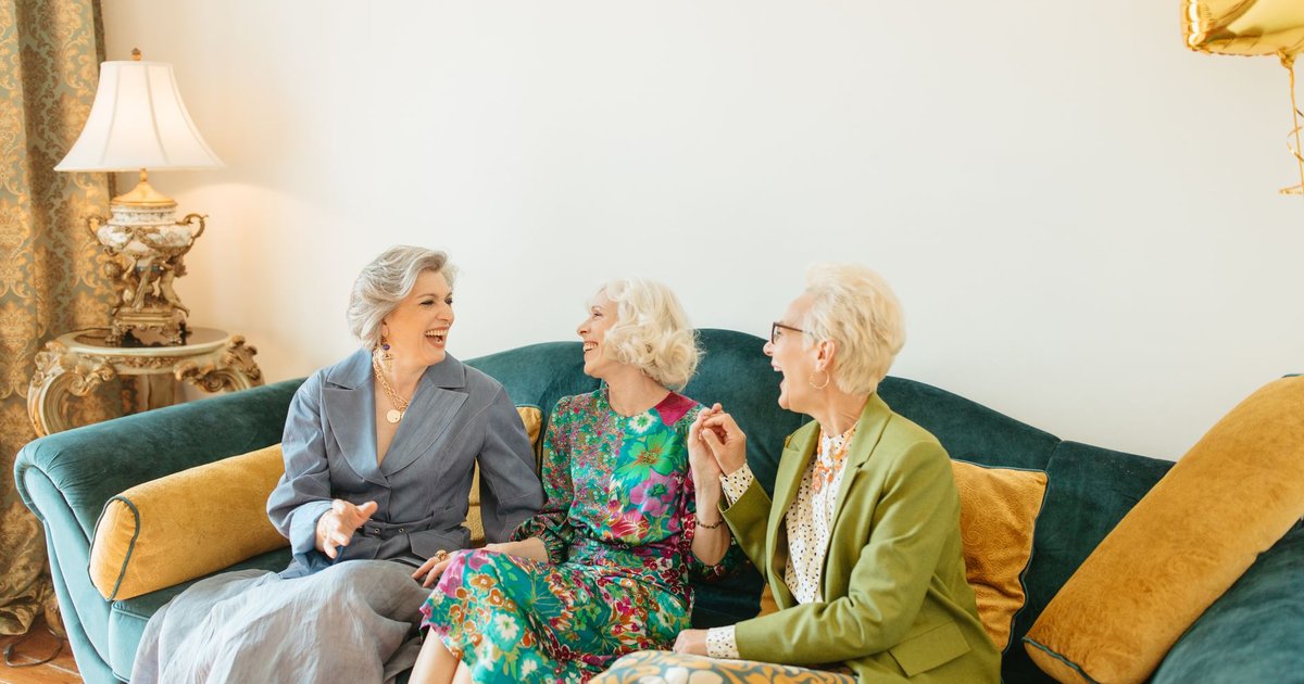 Three smiling older women laughing together on a sofa in a bright living room