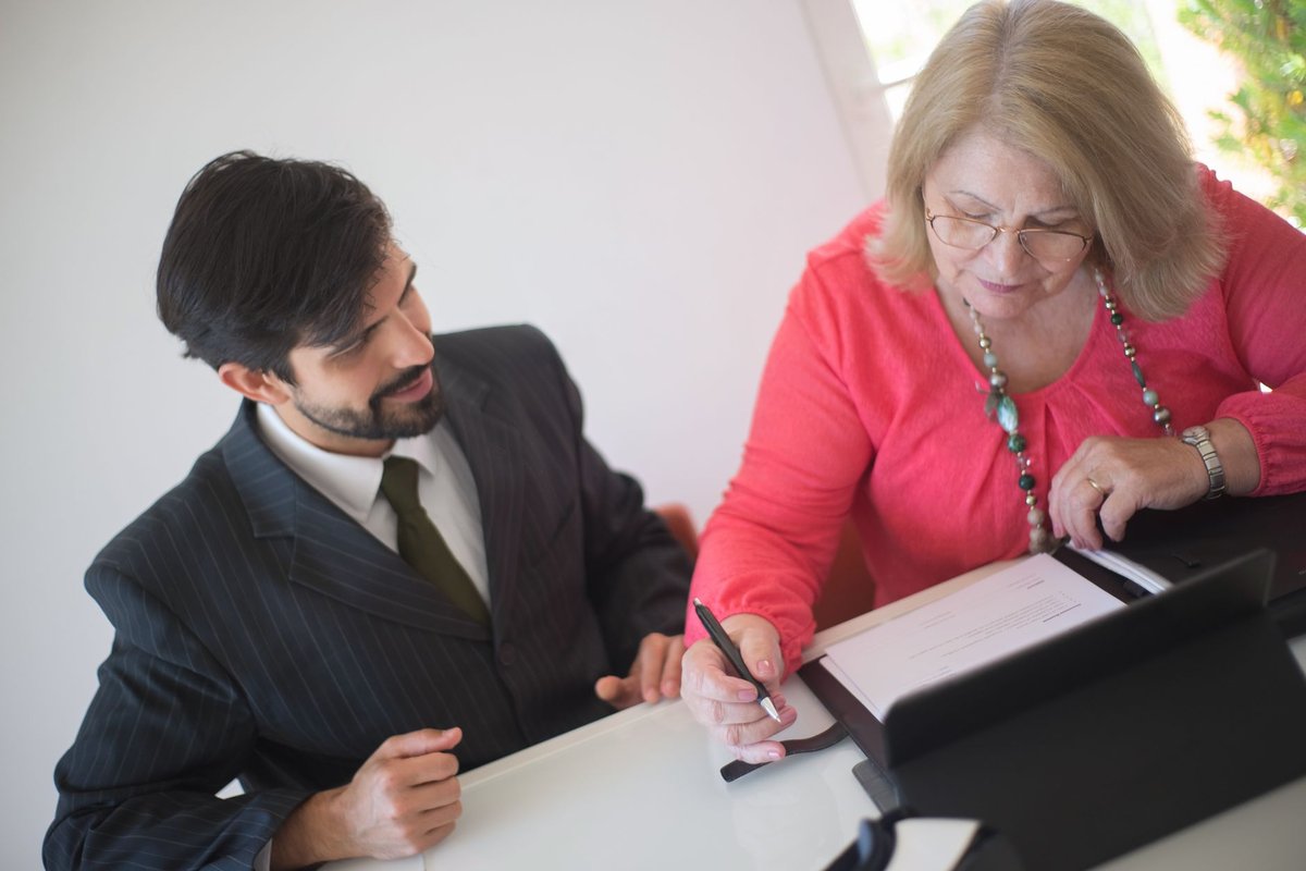 Older woman in a coral blouse signing paperwork while a man in a suit looks on