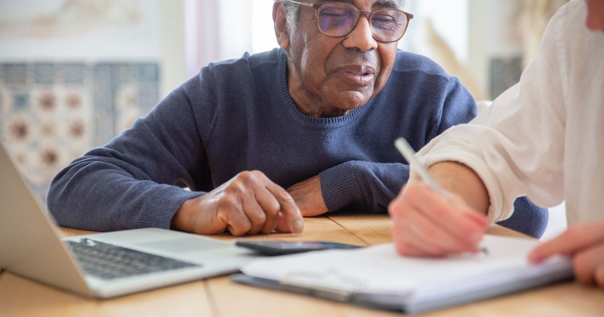 Older man in a navy sweater watches as papers are signed beside a laptop