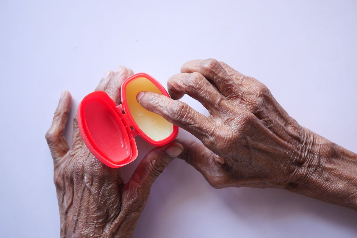 Older hands with deep wrinkles dipping a finger into a red tub of ointment