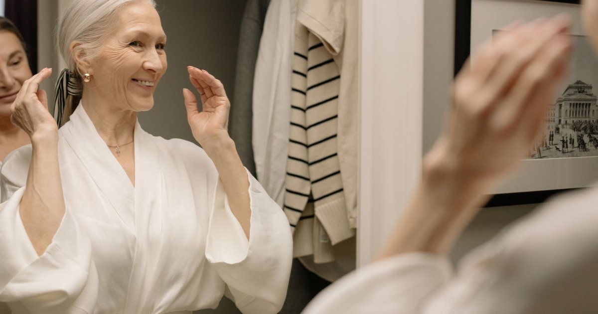 Smiling older woman with gray hair in a white robe looking at her reflection in a mirror
