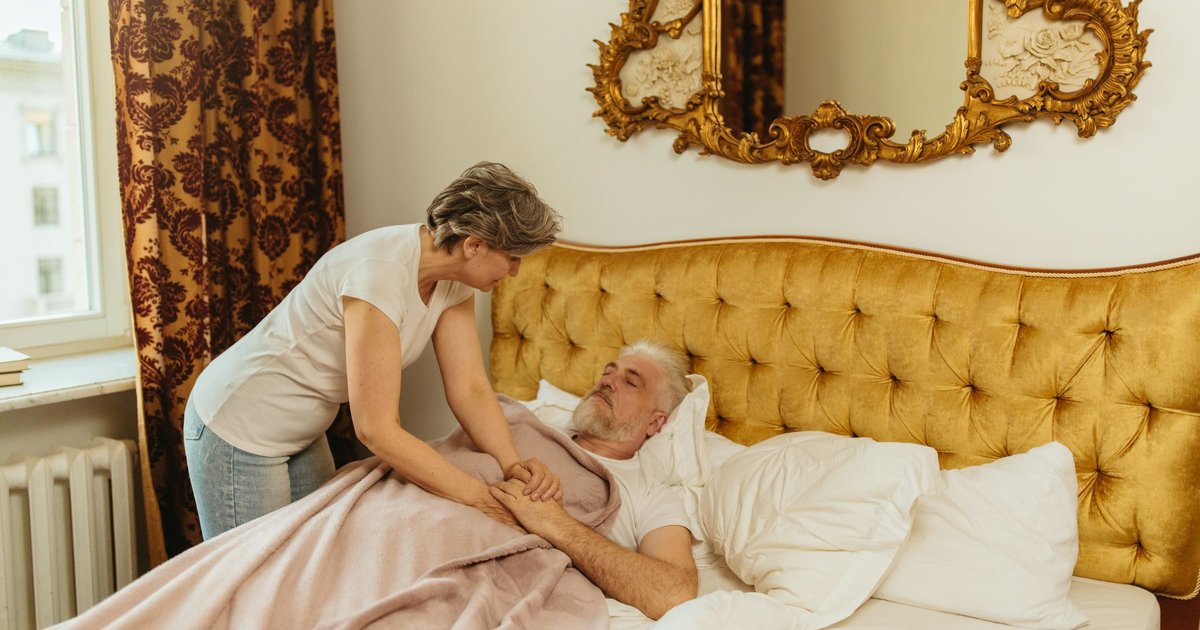 Woman gently holding the hands of an older man resting in bed at home