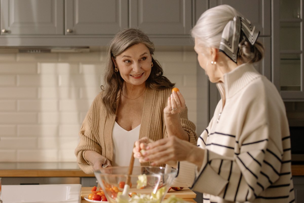 Two older women smiling and preparing a fresh salad together in a bright kitchen