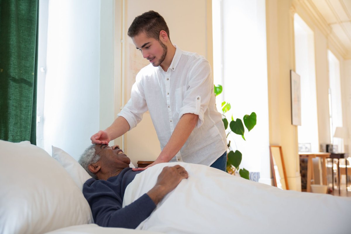 Caregiver leaning over an older man resting in bed, checking on his comfort