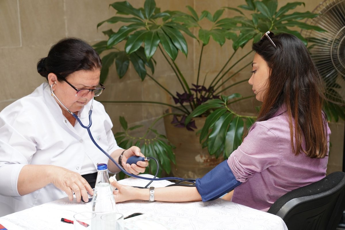 Nurse in a white coat taking a woman's blood pressure outdoors beside green plants