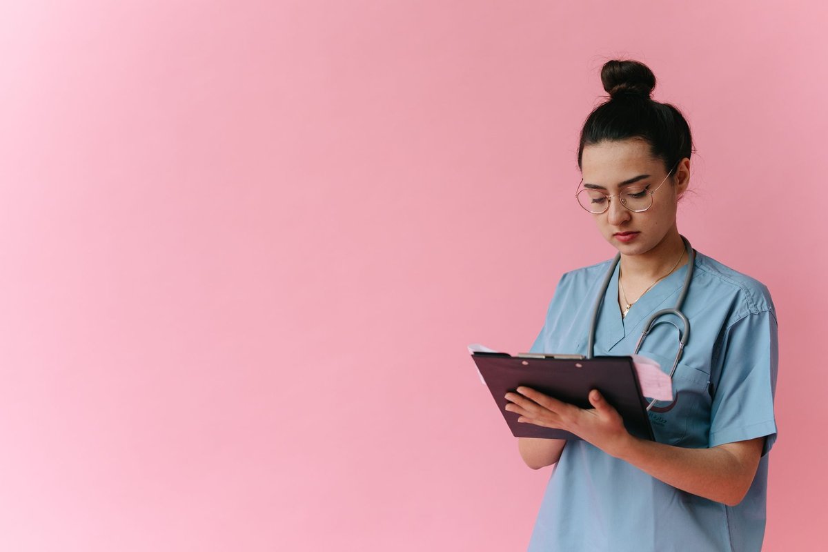 Nurse in light blue scrubs with a stethoscope writing on a clipboard against a pink wall