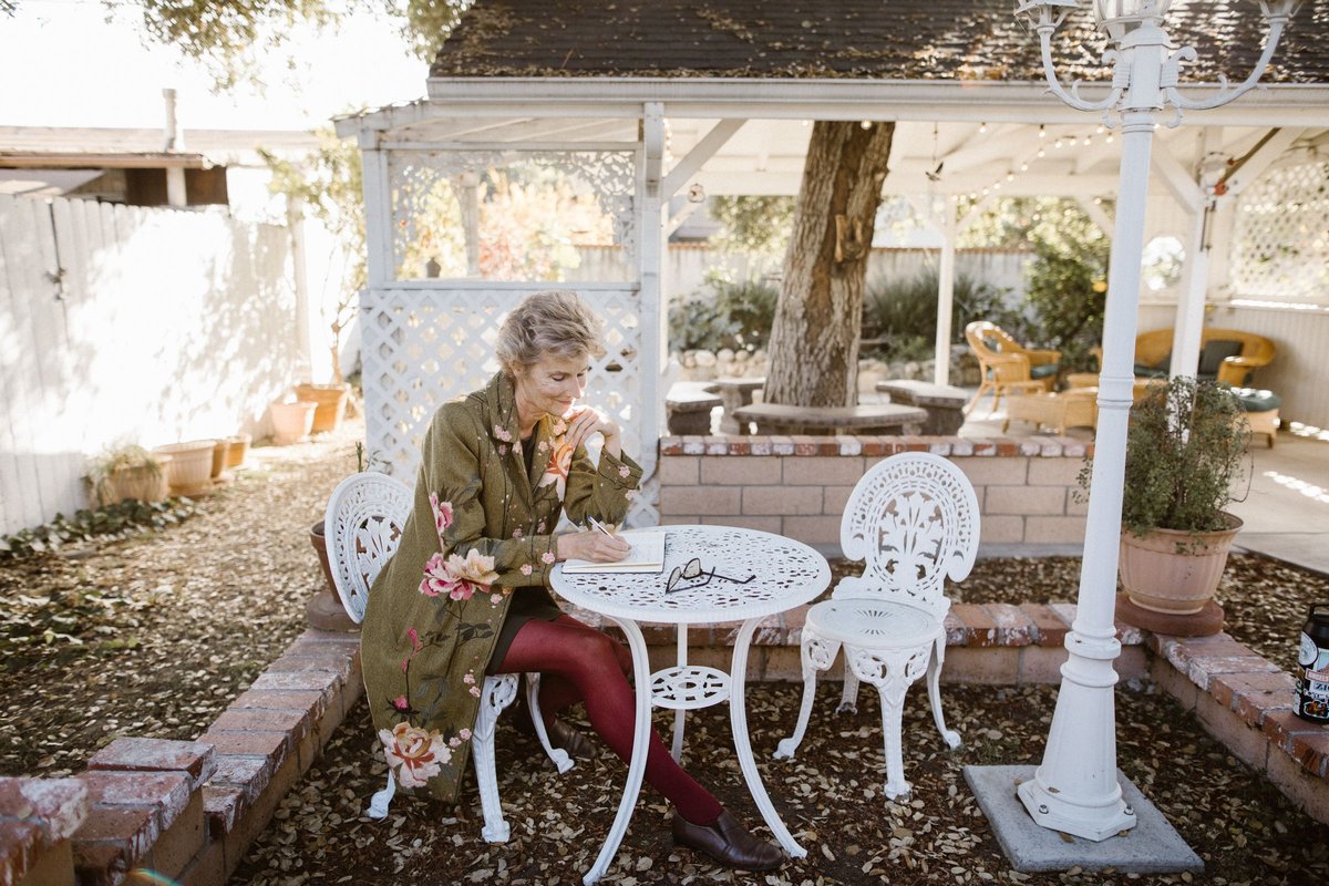 Older woman in a flowered coat writing in a notebook at a white garden table