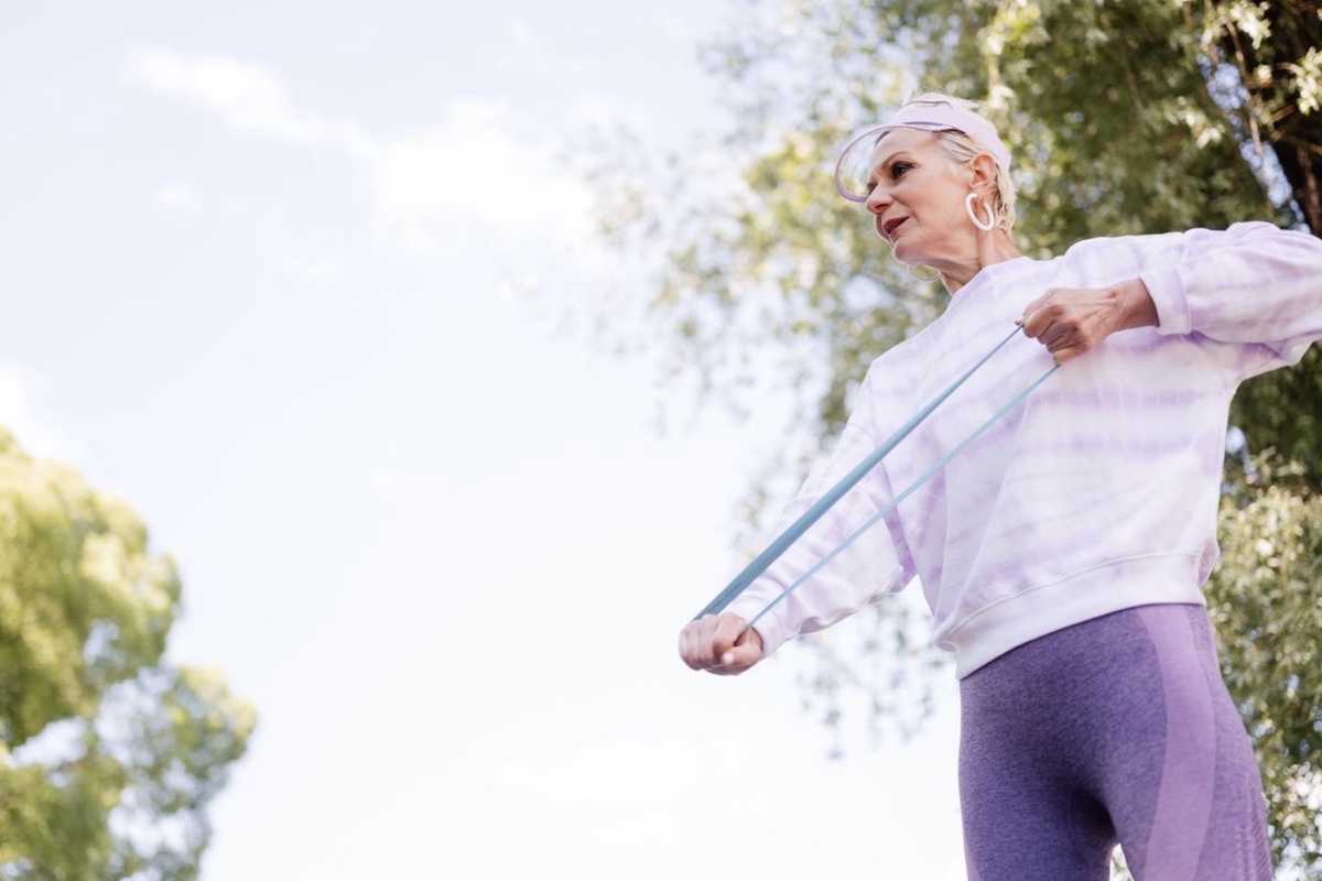 Older woman in lavender sportswear stretching an exercise band outdoors under trees