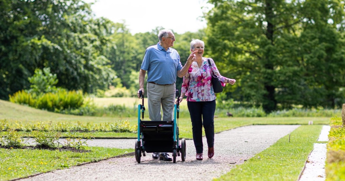 Older couple laughing as they walk along a sunny garden path with a rollator