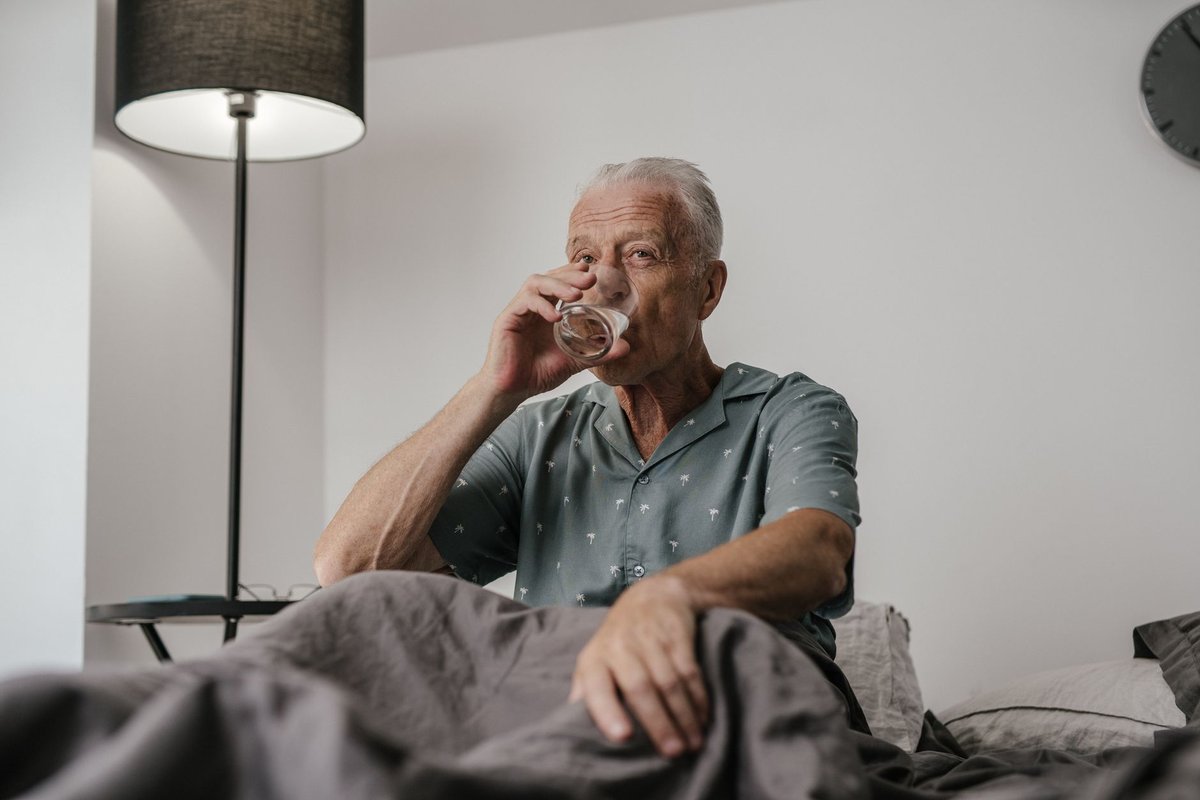 Older man with gray hair sitting up in bed drinking a glass of water