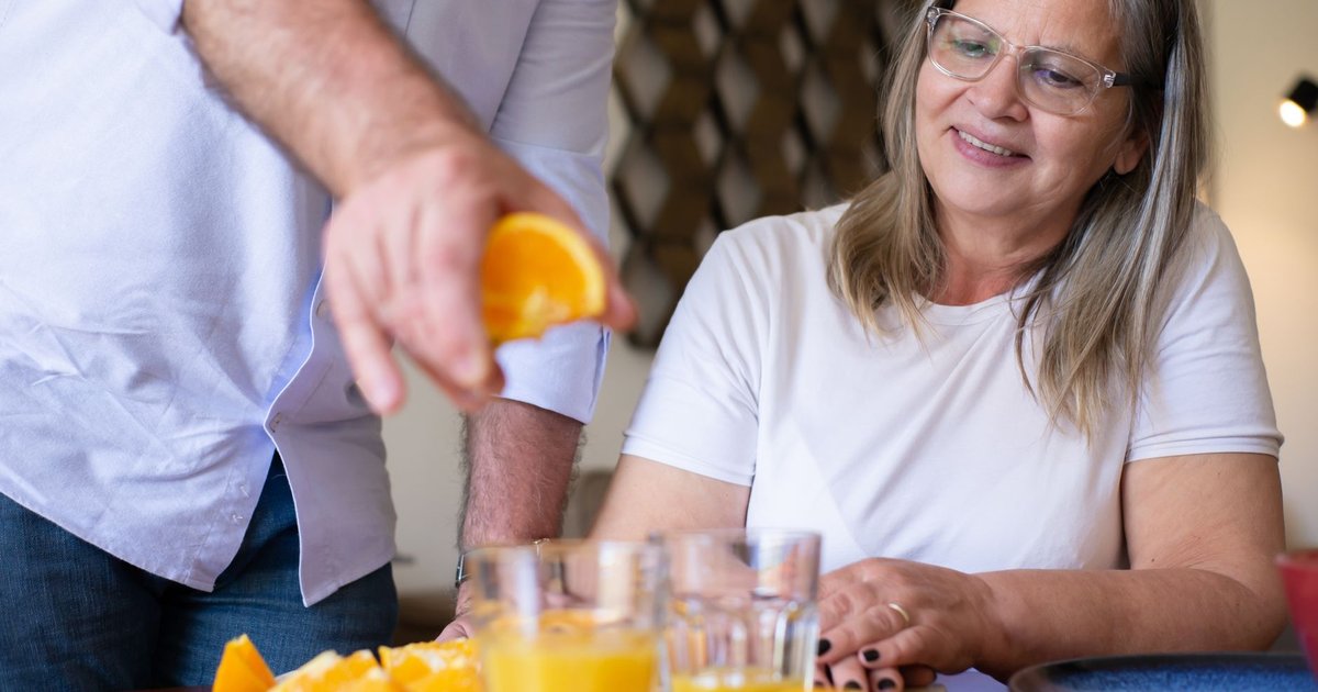 Smiling older woman watching glasses of fresh orange juice being poured at a kitchen table