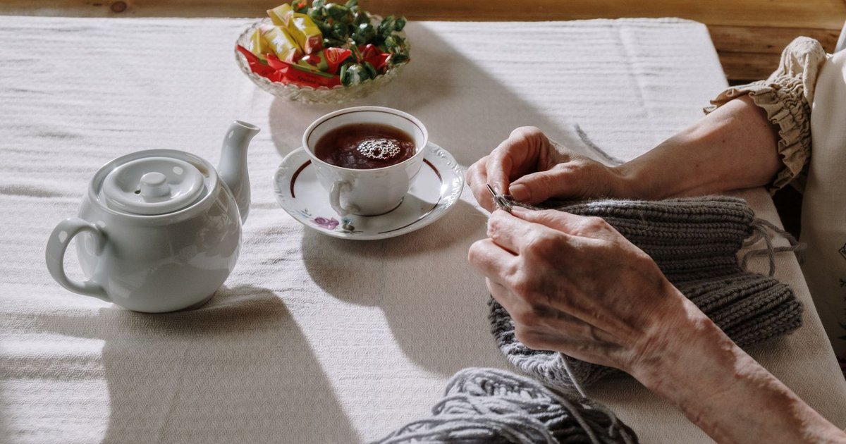 Older woman's hands knitting beside a cup of tea and a teapot on a table