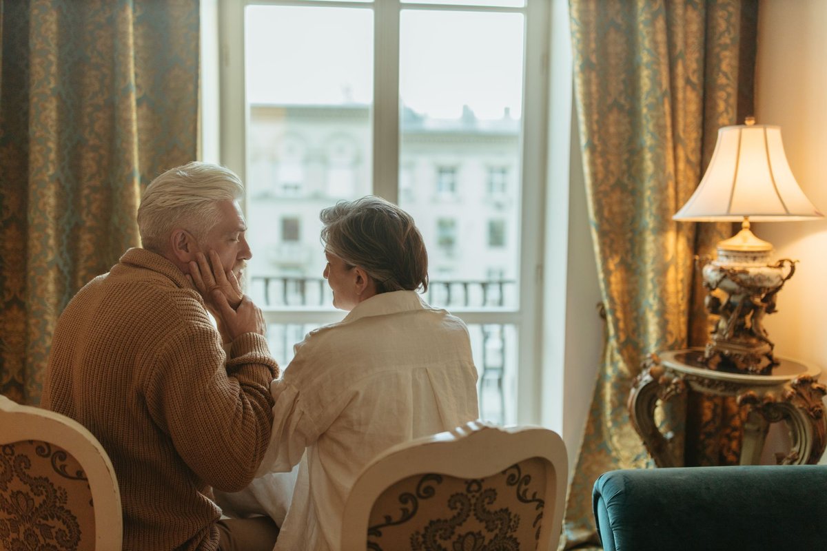 Older couple sitting close together by a bright window in a warmly lit living room