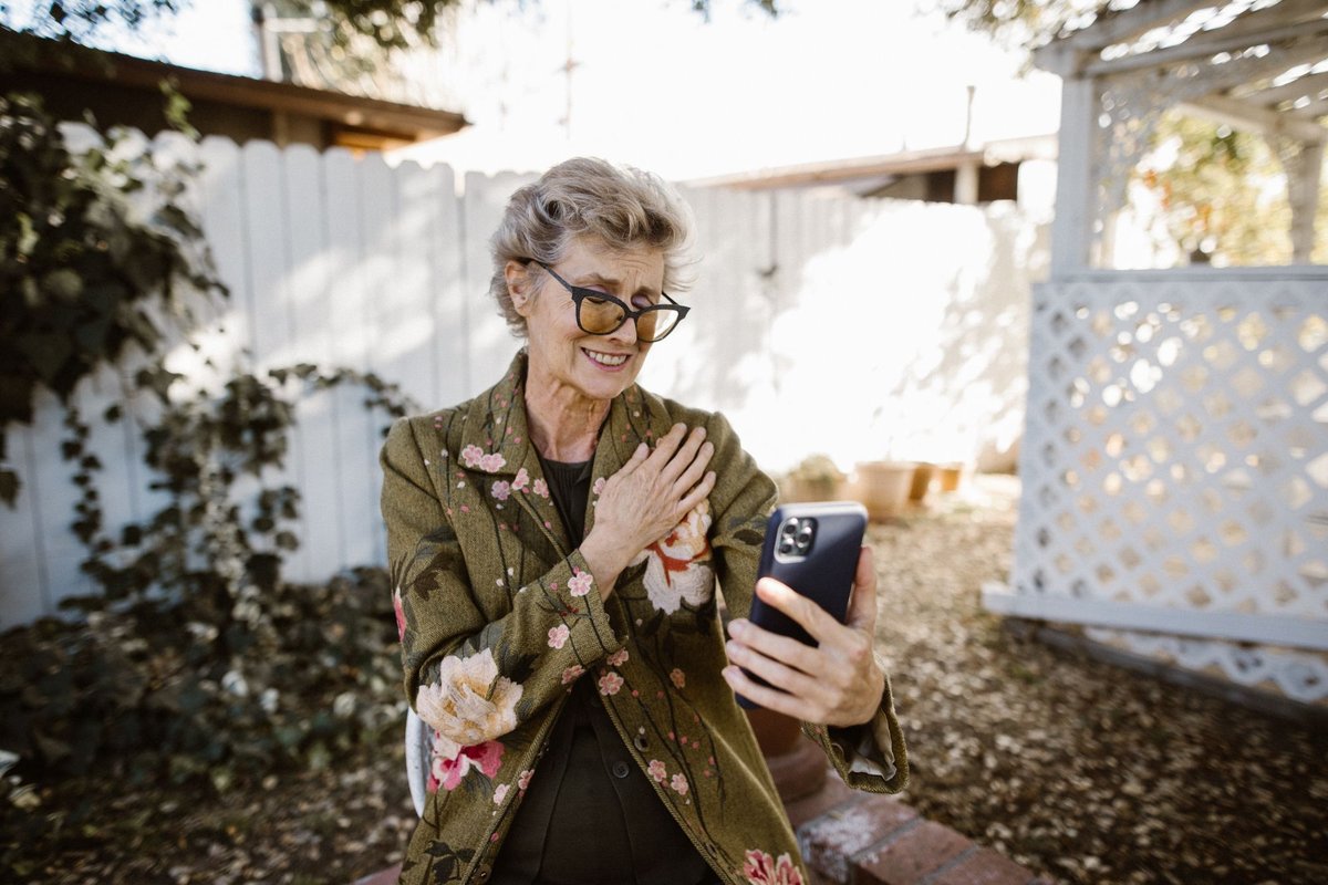 Smiling older woman with glasses looking at a smartphone during a video call in her backyard
