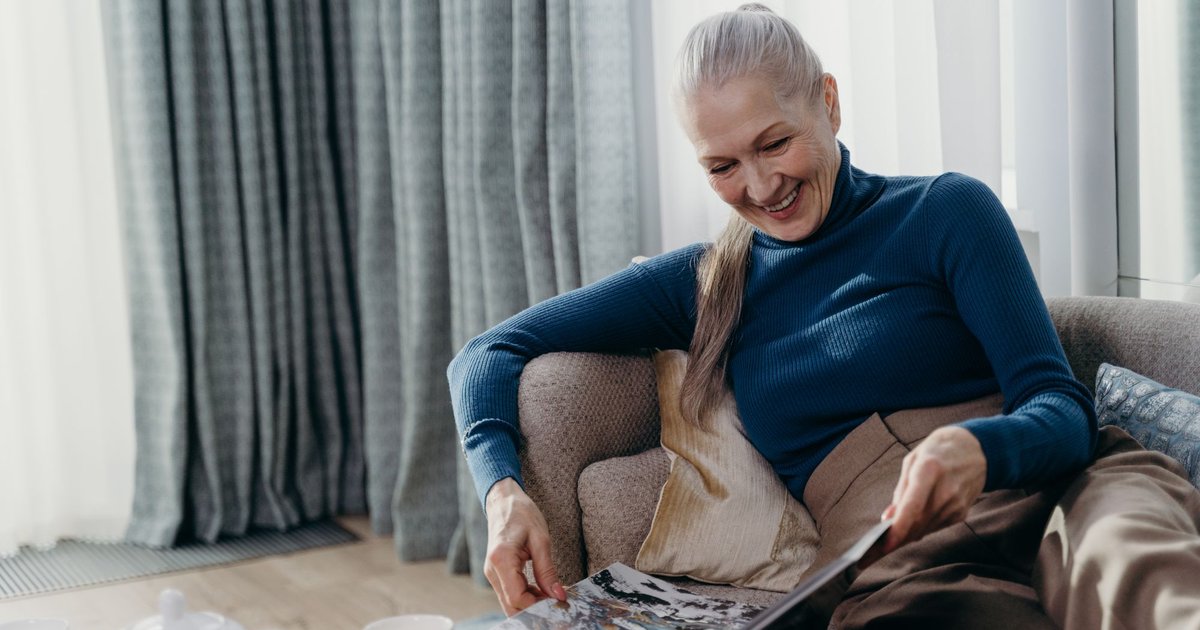 Older woman with a gray ponytail smiling as she looks through a magazine on a sofa