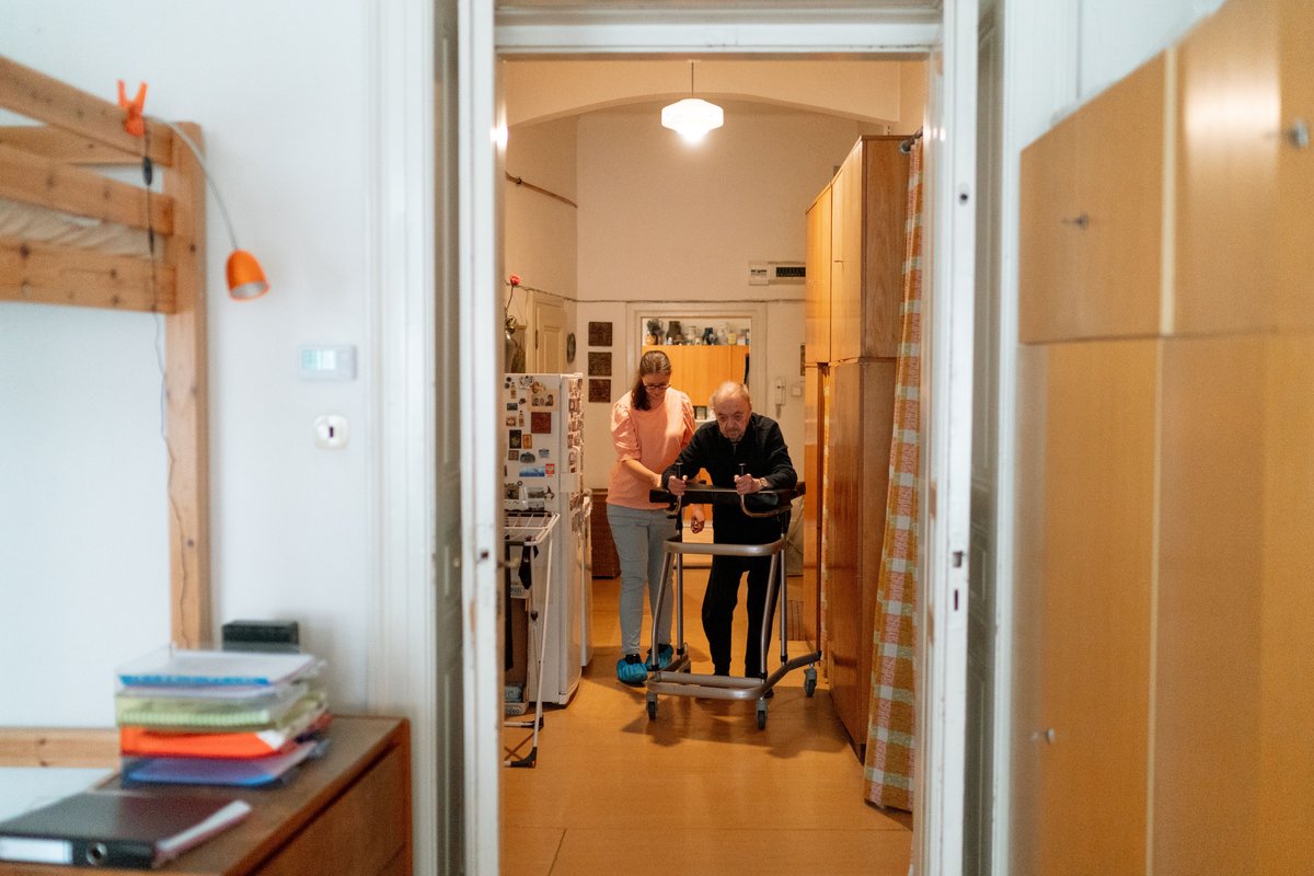 Home care aide walking beside an older man who uses a walker in a hallway