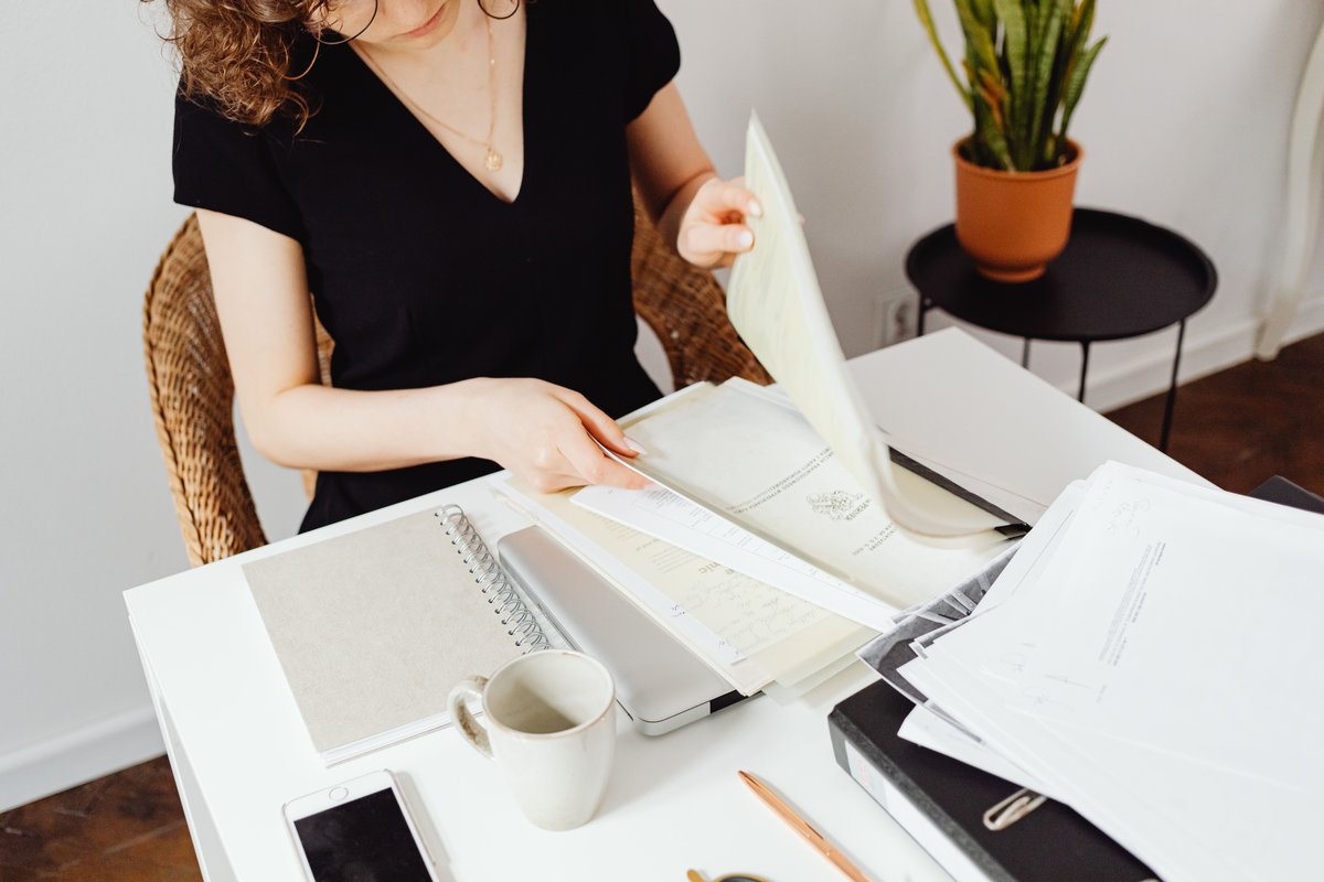 Woman sorting certificates and paperwork at a home desk with a notebook and coffee
