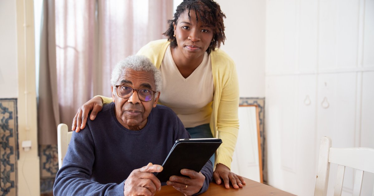 Home care aide standing behind an older man at a table while he holds a tablet