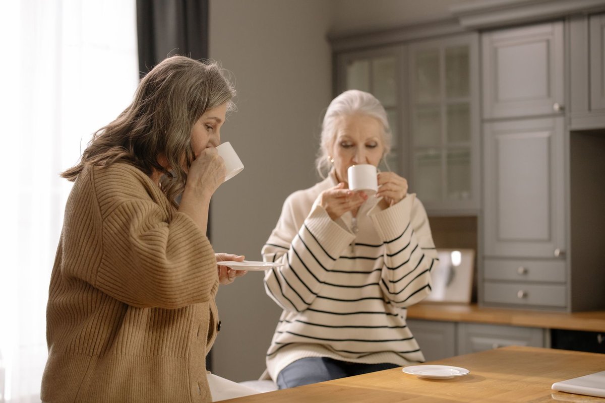 Two older women sipping from white cups at a kitchen counter in soft daylight