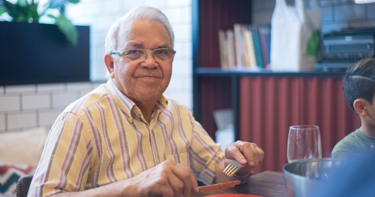 Smiling older man at a dining table holding a fork, with a glass and a child nearby
