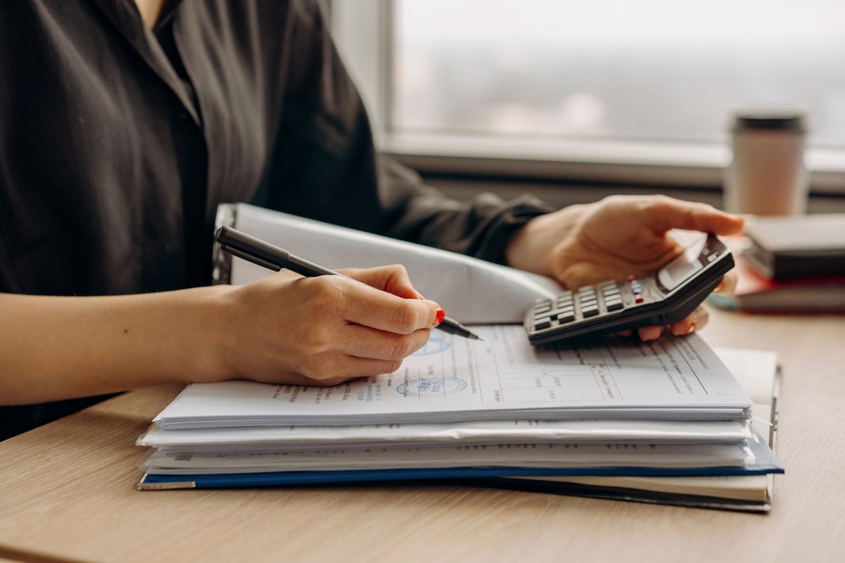 Hands hold a pen and a calculator over a stack of paper forms on a desk