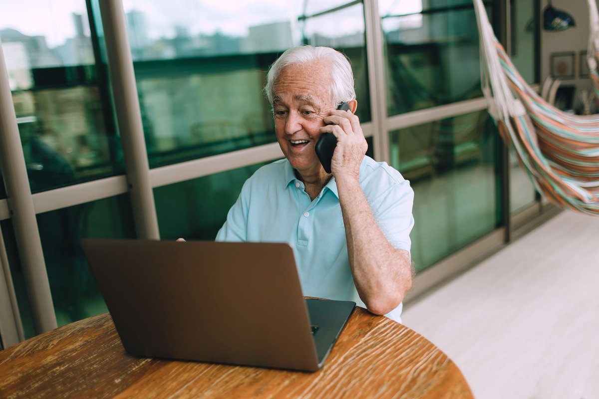 Smiling older man in a light blue shirt talks on the phone beside a laptop