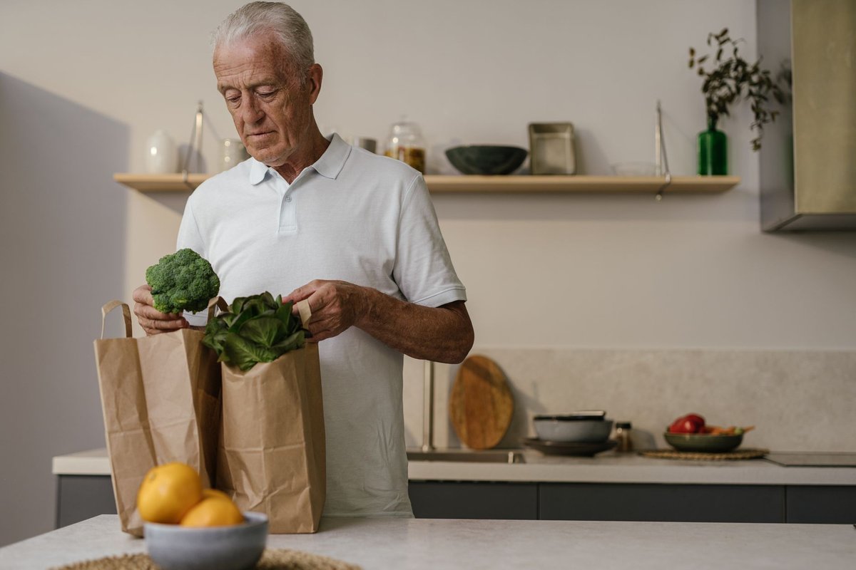Older man unpacking fresh broccoli and greens from grocery bags in his home kitchen