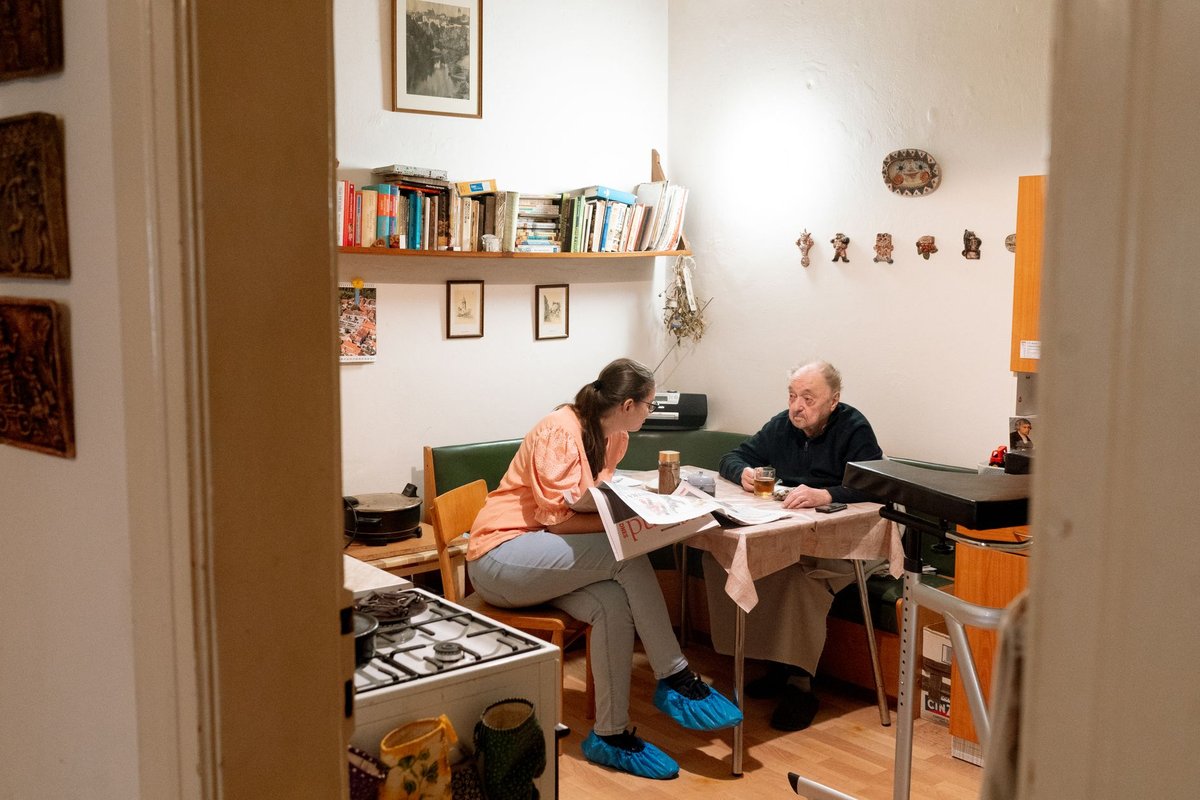 Home aide and an older man talking at a kitchen table over a newspaper and tea
