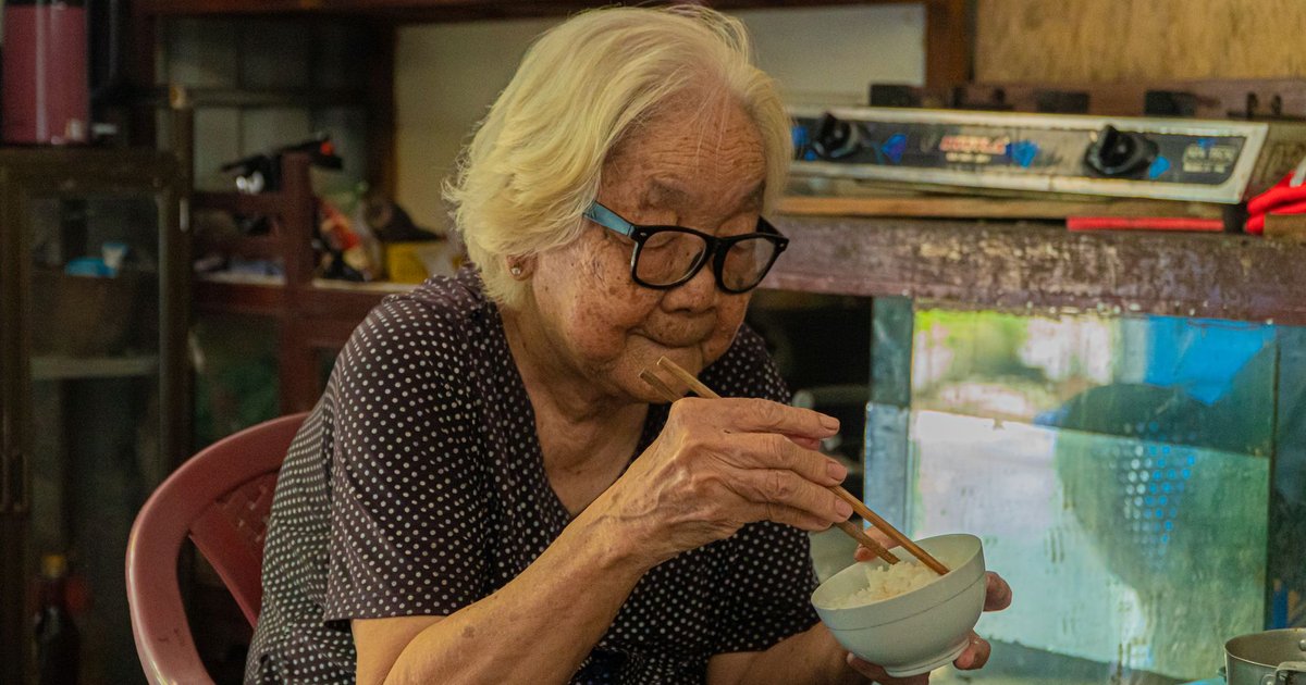 Older woman with white hair and glasses eats a bowl of rice with chopsticks