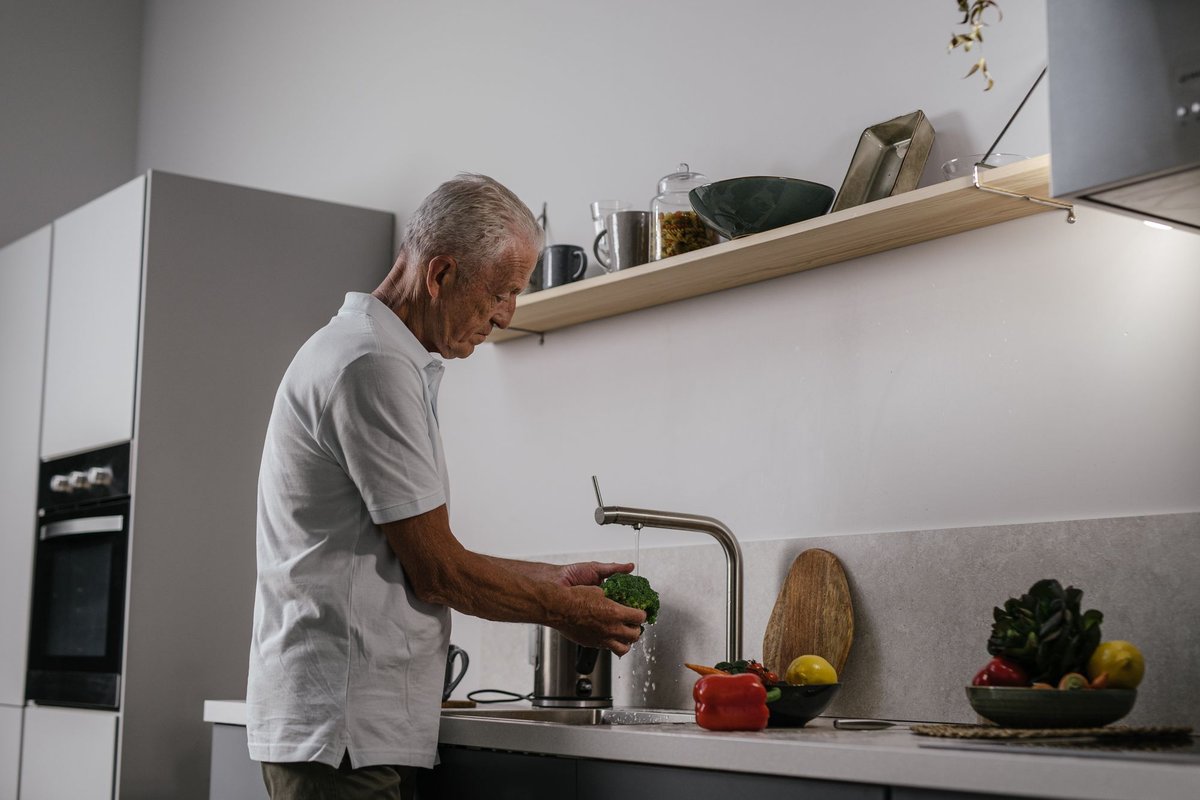 Older man in a bright kitchen rinsing fresh broccoli at the sink