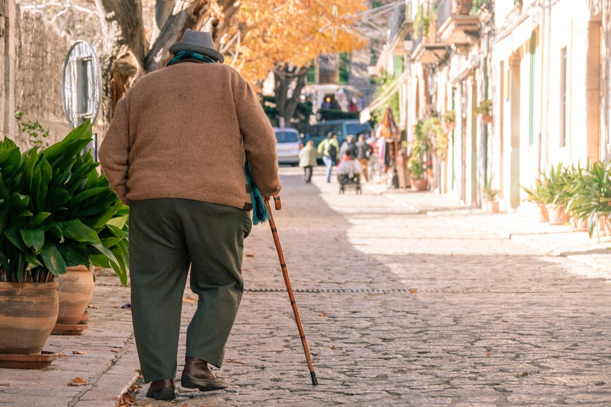 Older man in a hat and brown sweater walking with a wooden cane along a sunny cobbled street