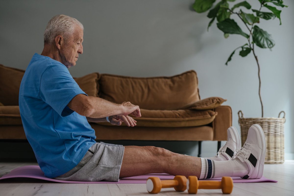 Older man in a blue shirt sitting on an exercise mat at home with hand weights beside him