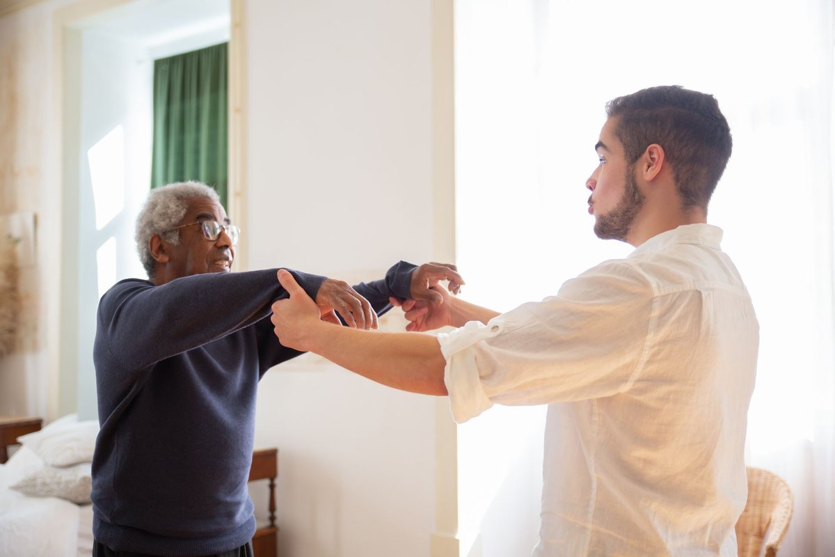 Caregiver helping an older man with gentle arm stretches in a bright bedroom