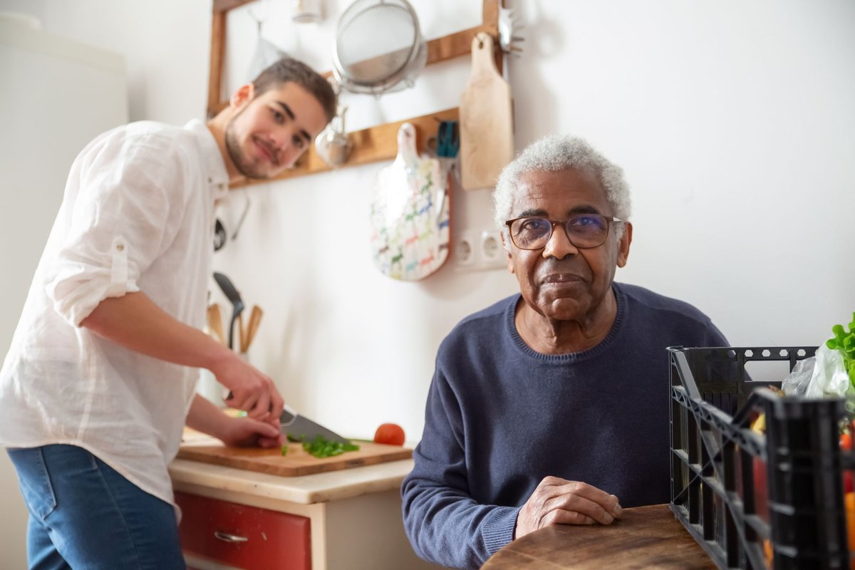 Young man chopping vegetables in a home kitchen while an older man sits nearby