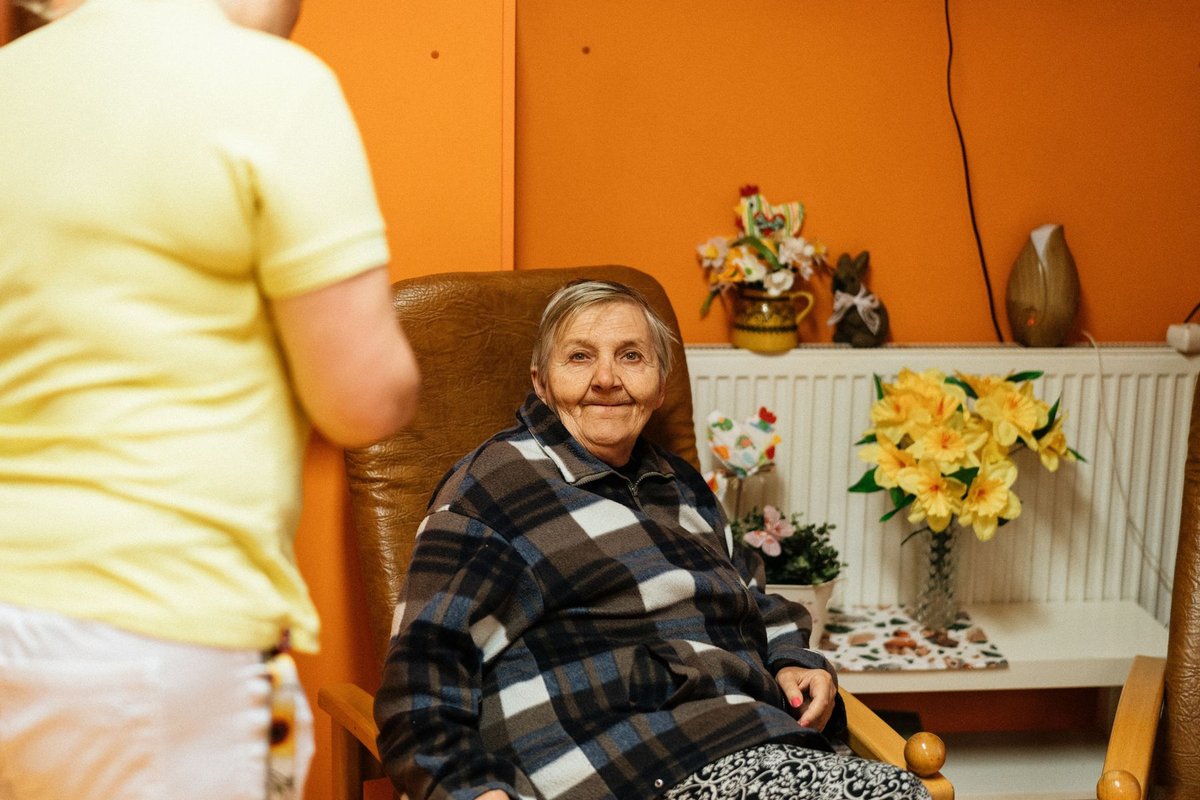 Older woman in an armchair smiling at a caregiver in a bright orange room