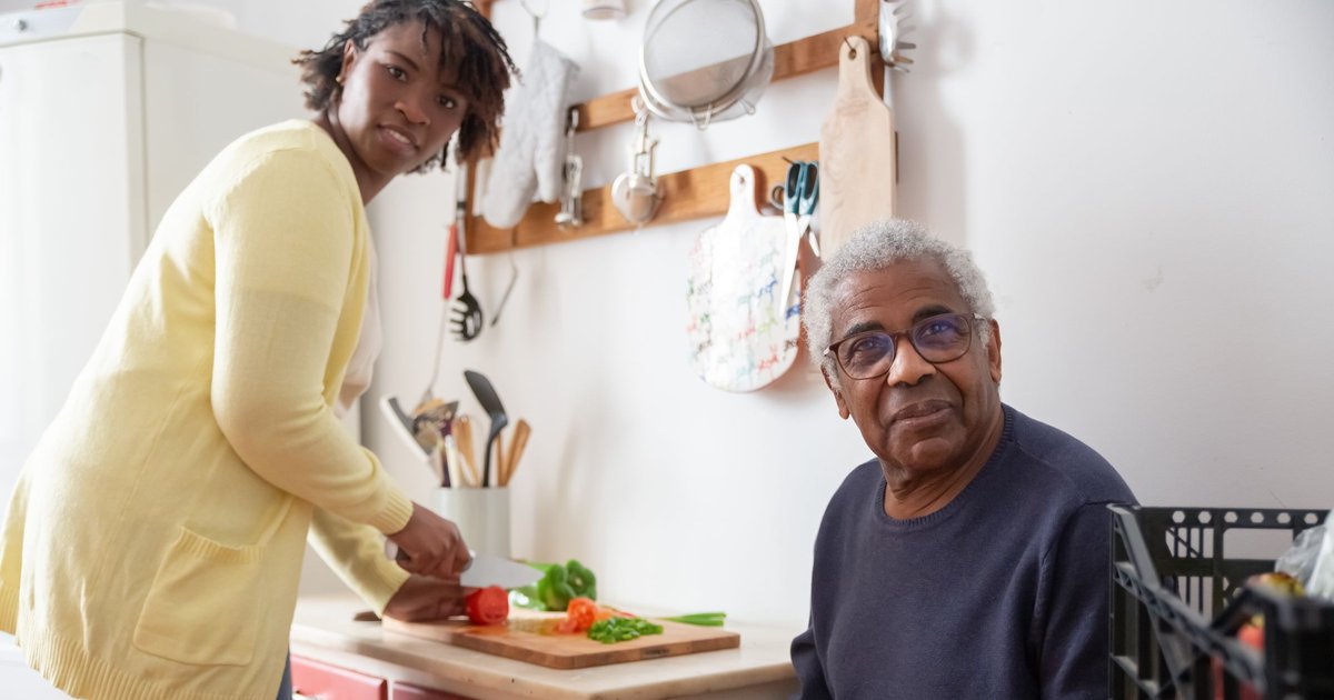 Woman in a yellow cardigan preparing vegetables in a kitchen next to a seated older man