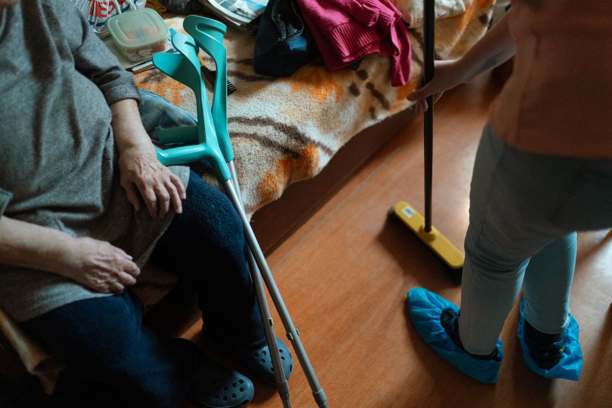 Seated older woman with crutches beside her while a helper sweeps the floor