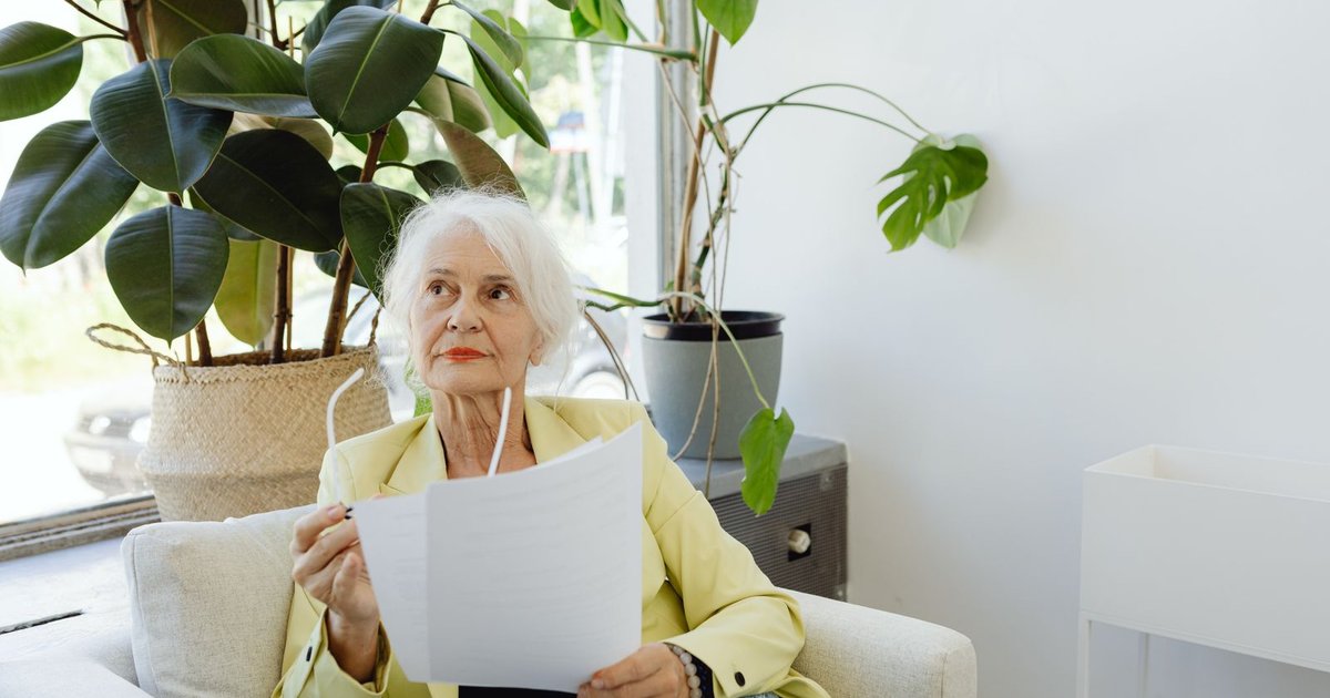 Older woman with white hair holding papers in an armchair near a bright window