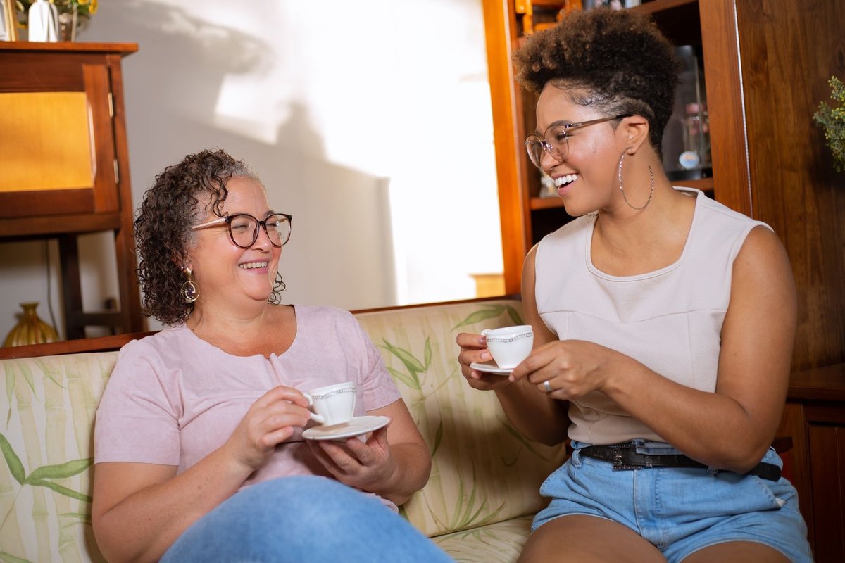 Two women laughing together over cups of tea on a sofa at home