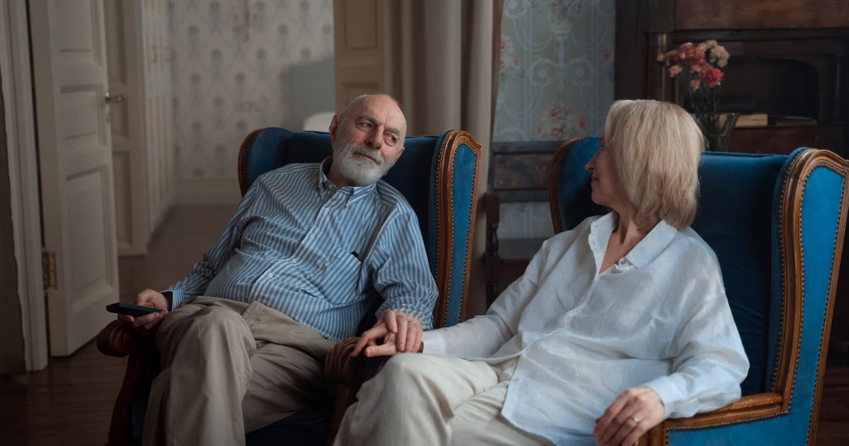 Elderly couple sitting side by side in armchairs at home, holding hands