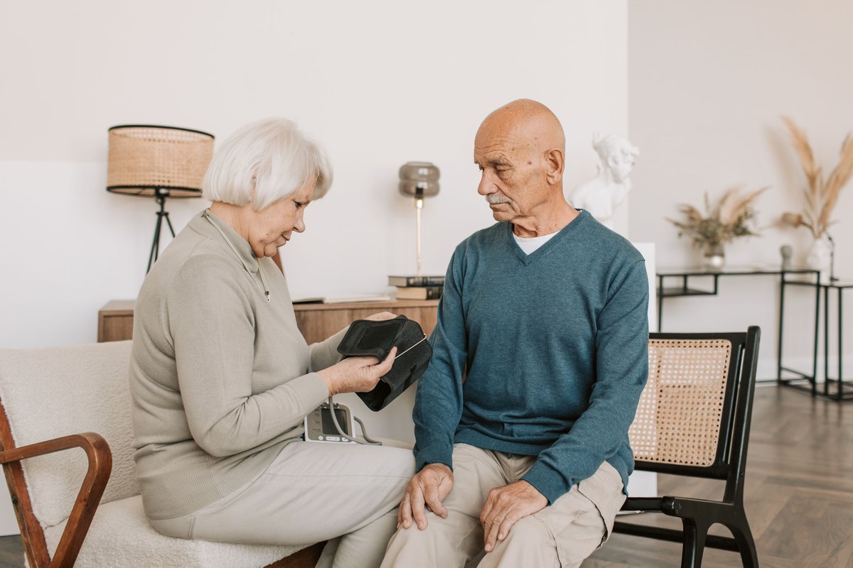 Older woman holding a blood pressure cuff next to an older man seated beside her at home