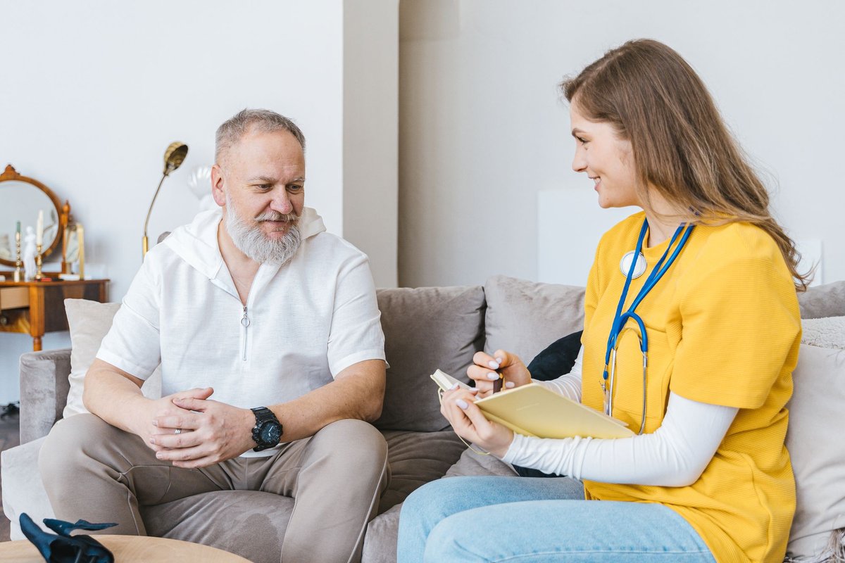 Nurse in a yellow top takes notes while talking with a man on a couch